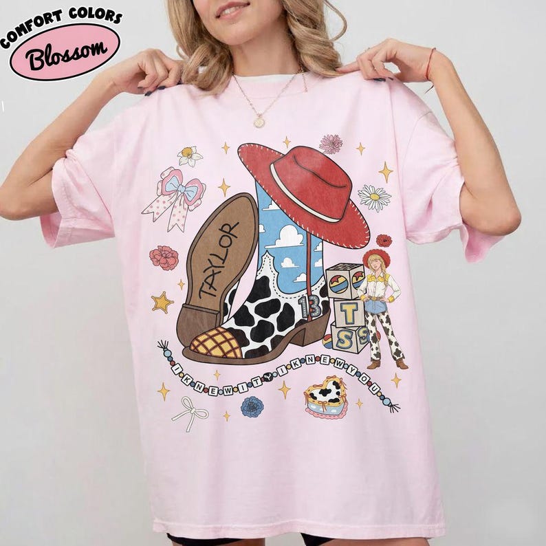 May include: A light pink t-shirt featuring a graphic print of a cowboy boot with a cow print pattern, a red hat, a character, and blocks with the letters T, S, and 13. The text "TAYLOR" and "I KNEW IT Y I KNEW Y OU" are printed on the shirt.
