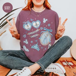May include: Berry-colored t-shirt featuring Disney Elsa graphics. The design includes images of Olaf, Elsa, snowflakes, and a crown. The text "Disney Elsa" is also visible. The shirt is a comfortable, casual style.