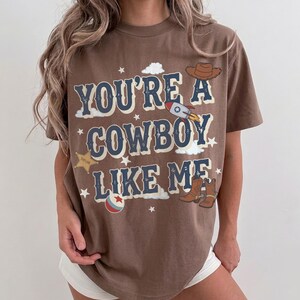 May include: A brown short-sleeve t-shirt featuring the text "YOU'RE A COWBOY LIKE ME" in dark blue letters. The graphic includes illustrations of a brown cowboy hat, a rocket ship, a gold sheriff star, a pair of brown boots, and small white stars.