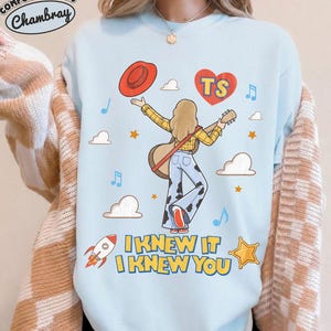 Puede incluir: Una sudadera azul claro de cuello redondo con un gr&aacute;fico de una persona de cabello rubio sosteniendo una guitarra, vistiendo una camisa de cuadros amarilla y pantalones con estampado de vaca. El dise&ntilde;o incluye un sombrero rojo, un coraz&oacute;n con TS, nubes, estrellas, un cohete y el texto I KNEW IT I KNEW YOU.
