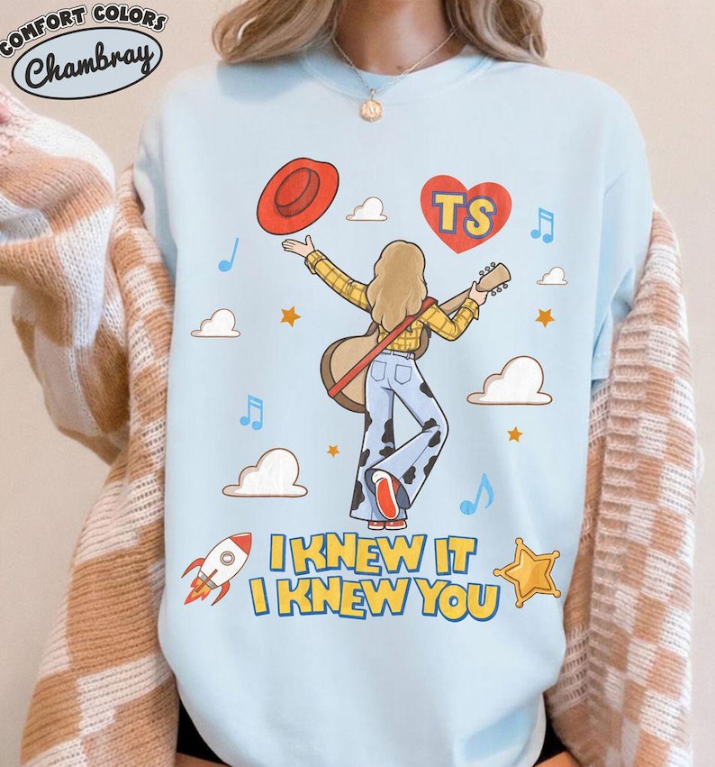 Puede incluir: Una sudadera azul claro de cuello redondo con un gr&aacute;fico de una persona de cabello rubio sosteniendo una guitarra, vistiendo una camisa de cuadros amarilla y pantalones con estampado de vaca. El dise&ntilde;o incluye un sombrero rojo, un coraz&oacute;n con TS, nubes, estrellas, un cohete y el texto I KNEW IT I KNEW YOU.