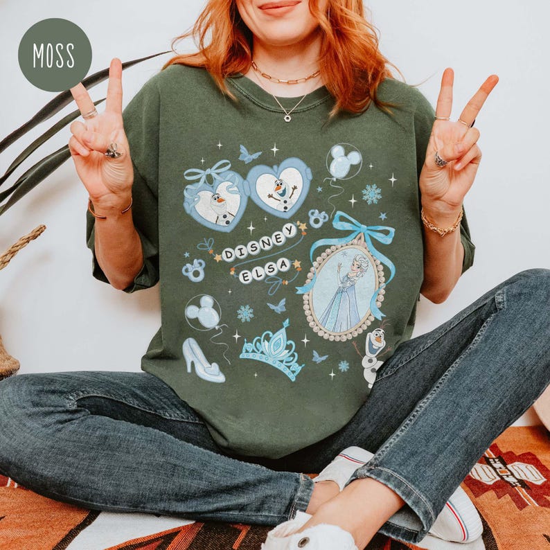 May include: Moss green graphic t-shirt featuring Disney's Elsa. The design includes images of Olaf, Elsa, snowflakes, a crown, and a high heel. The words "Disney Elsa" are in a decorative font. The shirt is a relaxed fit.
