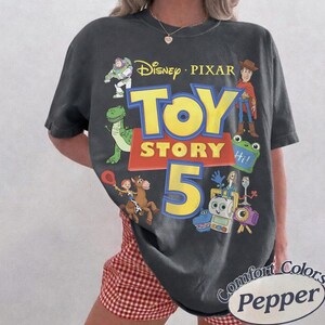 Puede incluir: Una camiseta de manga corta gris carb&oacute;n con un estampado gr&aacute;fico que incluye el texto Disney Pixar Toy Story 5. El dise&ntilde;o presenta ilustraciones de Buzz Lightyear, Woody, Rex, Jessie, Bullseye, Forky y otros personajes en colores vivos.