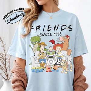 K&ouml;nnte beinhalten: Hellblaues T-Shirt mit dem Wort "FRIENDS" in Schwarz und "SINCE 1995" darunter. Das Shirt zeigt eine Grafik von Toy Story-Charakteren. Das Shirt besteht aus Comfort Colors Chambray-Material.