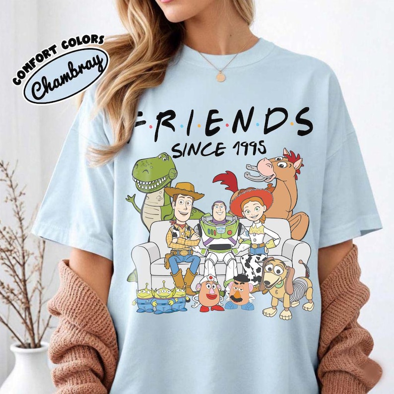 K&ouml;nnte beinhalten: Hellblaues T-Shirt mit dem Wort "FRIENDS" in Schwarz und "SINCE 1995" darunter. Das Shirt zeigt eine Grafik von Toy Story-Charakteren. Das Shirt besteht aus Comfort Colors Chambray-Material.