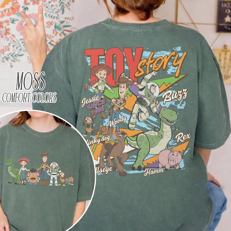 Może przedstawiać: Koszulka w kolorze mchu z nadrukiem postaci z Toy Story. Projekt zawiera Jessie, Woody, Buzz, Rex i Hamm, z napisem "Toy Story" i "Since 1995".