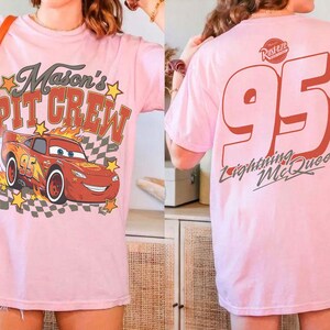 Op de afbeelding: Een lichtroze t-shirt met een afbeelding op de voorkant van een rode raceauto met vlammen, een dambordpatroon, gele sterren en de tekst Mason's PIT CREW. De achterkant toont het nummer 95, het Rust-eze logo en de tekst Lightning McQueen.