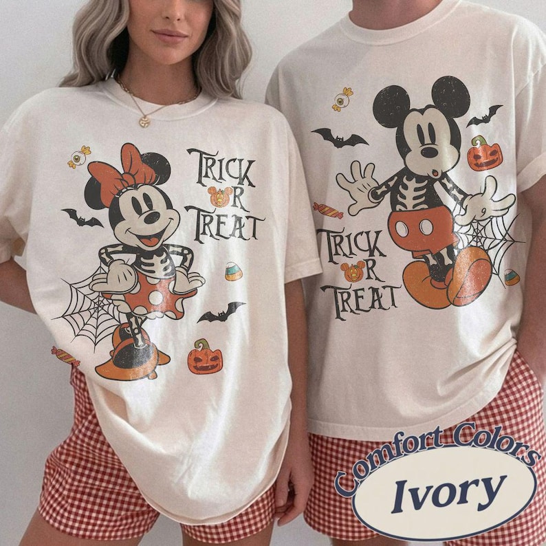 May include: Two ivory t-shirts featuring Halloween-themed graphics of Mickey and Minnie Mouse in skeleton costumes. The designs include the text "Trick or Treat," spiderwebs, bats, pumpkins, and candy corn, printed in black, orange, and white colors.