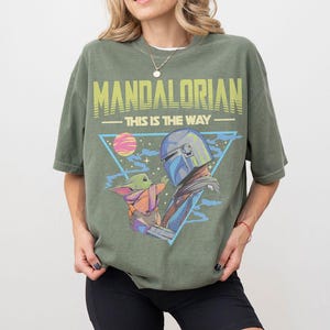 Może przedstawiać: Oliwkowozielony t-shirt z kr&oacute;tkim rękawem z nadrukiem graficznym przedstawiającym Mandalorianina i Grogu. Napis głosi MANDALORIAN oraz THIS IS THE WAY w kolorze ż&oacute;łtym i jasnym zielonym. Wz&oacute;r zawiera niebieski tr&oacute;jkąt, r&oacute;żową planetę i gwiazdy.