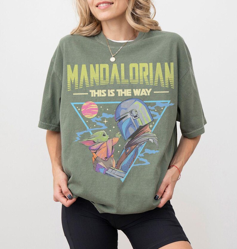 Może przedstawiać: Oliwkowozielony t-shirt z kr&oacute;tkim rękawem z nadrukiem graficznym przedstawiającym Mandalorianina i Grogu. Napis głosi MANDALORIAN oraz THIS IS THE WAY w kolorze ż&oacute;łtym i jasnym zielonym. Wz&oacute;r zawiera niebieski tr&oacute;jkąt, r&oacute;żową planetę i gwiazdy.