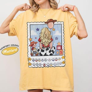May include: A mustard yellow t-shirt featuring a graphic print of a blonde person seen from behind, a small cowgirl doll, a cowboy hat balloon, and alphabet blocks. The text reads "EVERY ERA LED TO YOU" inside a decorative postage stamp border.