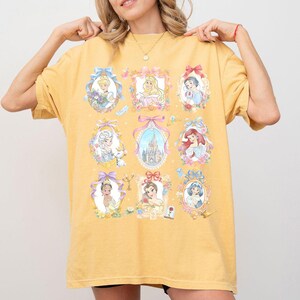 May include: Mustard yellow t-shirt featuring framed portraits of Disney princesses. Each portrait is surrounded by floral and bow accents. The shirt's design includes images of Cinderella, Snow White, Ariel, and other beloved characters.