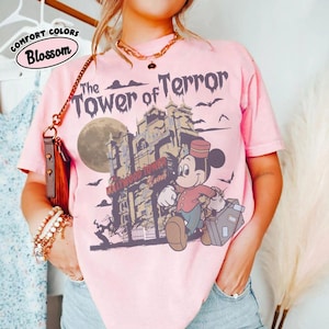 Puede incluir: Camiseta rosa claro con un gr&aacute;fico de Mickey Mouse como botones frente al Hollywood Tower Hotel. El texto "The Tower of Terror" est&aacute; encima de la imagen, con las palabras "Comfort Colors Blossom" en un c&iacute;rculo.