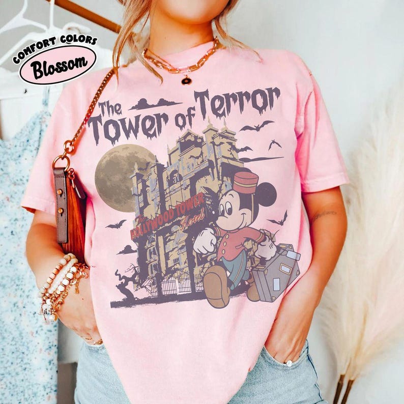 Puede incluir: Camiseta rosa claro con un gr&aacute;fico de Mickey Mouse como botones frente al Hollywood Tower Hotel. El texto "The Tower of Terror" est&aacute; encima de la imagen, con las palabras "Comfort Colors Blossom" en un c&iacute;rculo.