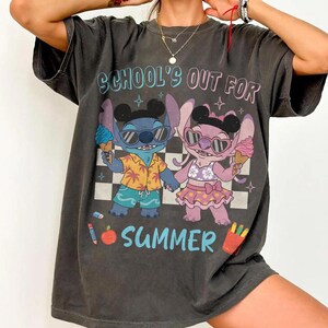 May include: Dark gray t-shirt featuring a cartoon illustration of two characters holding ice cream cones. The text "SCHOOL'S OUT FOR SUMMER" is printed above the characters in blue and pink. The characters are wearing sunglasses and summer attire.
