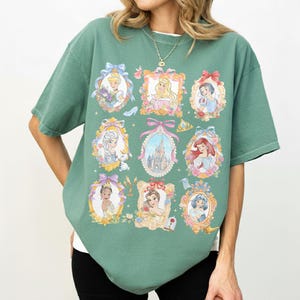 May include: Green t-shirt featuring framed portraits of animated princesses. The portraits are in ornate gold frames with floral and bow accents. The shirt has a crew neck and short sleeves.
