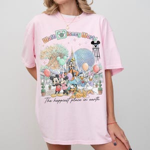 May include: Pink t-shirt featuring a colorful graphic of Disney World characters, including Mickey and Minnie Mouse, Goofy, Donald and Daisy Duck. The text "Walt Disney World" and "The happiest place in earth" are also visible.