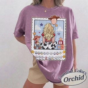 May include: A purple short-sleeve t-shirt featuring a graphic print of a blonde person with a cowboy hat balloon, a small cowgirl doll, and alphabet blocks. The text reads EVERY ERA LED TO YOU. The design is framed by a scalloped border with stars.