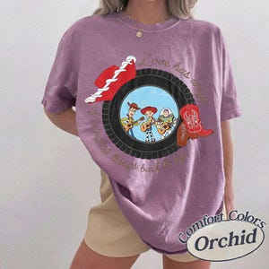 May include: A purple short-sleeve t-shirt featuring a graphic of a black tire framing cartoon characters Woody, Jessie, and Buzz Lightyear. The design includes a red cowboy hat, a red cowboy boot, and the text Love has a way of bringing things back to life.