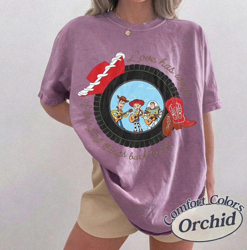 May include: A purple short-sleeve t-shirt featuring a graphic of a black tire framing cartoon characters Woody, Jessie, and Buzz Lightyear. The design includes a red cowboy hat, a red cowboy boot, and the text Love has a way of bringing things back to life.