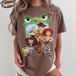 May include: A brown short-sleeve t-shirt featuring a graphic of Toy Story characters including Woody, Buzz Lightyear, Jessie, and Bullseye. The design includes the text Toy Story 5 In Theaters June 19 and a green frog frame. A Comfort Colors Espresso label is visible.