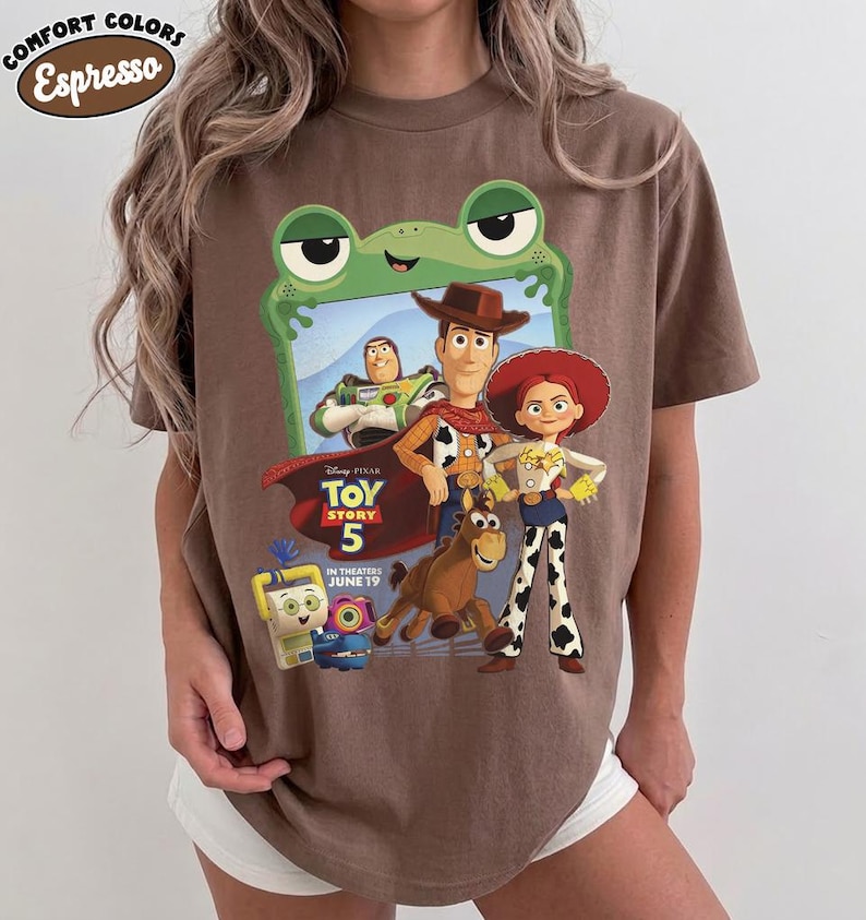 May include: A brown short-sleeve t-shirt featuring a graphic of Toy Story characters including Woody, Buzz Lightyear, Jessie, and Bullseye. The design includes the text Toy Story 5 In Theaters June 19 and a green frog frame. A Comfort Colors Espresso label is visible.
