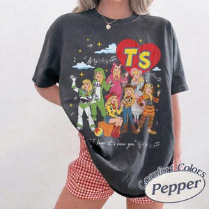 Puede incluir: Una camiseta gris carb&oacute;n de manga corta con un gr&aacute;fico de un gran coraz&oacute;n rojo con las letras TS. El dise&ntilde;o incluye personajes de dibujos animados con varios disfraces, notas musicales, estrellas y el texto I knew it, I knew you.