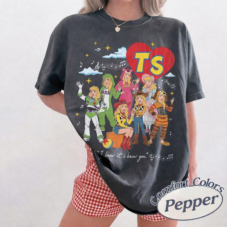 Puede incluir: Una camiseta gris carb&oacute;n de manga corta con un gr&aacute;fico de un gran coraz&oacute;n rojo con las letras TS. El dise&ntilde;o incluye personajes de dibujos animados con varios disfraces, notas musicales, estrellas y el texto I knew it, I knew you.