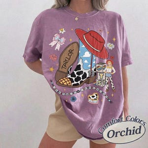 May include: A purple short-sleeve t-shirt featuring a graphic print of a cowboy boot with a cow print pattern, a red hat, a small figure, and blocks with the letters T, S, and the number 13. The text TAYLOR is on the boot sole.