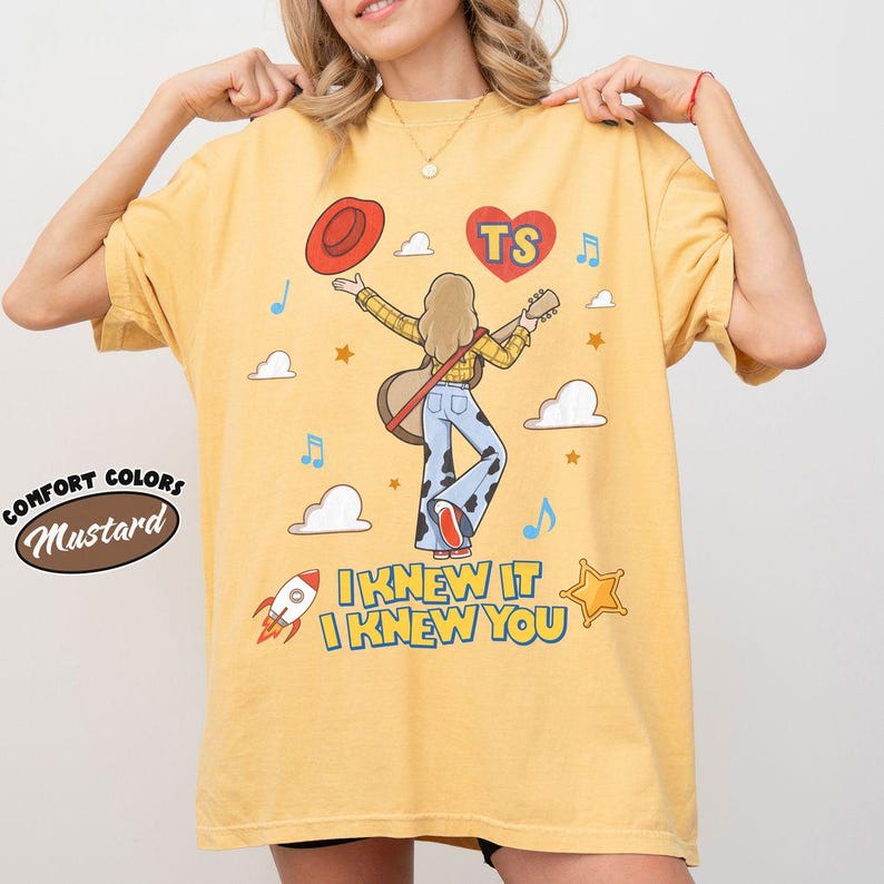 Puede incluir: Una camiseta color amarillo mostaza con un gr&aacute;fico de una persona de pelo largo que viste una camisa de cuadros amarilla y pantalones con estampado de vaca, sosteniendo una guitarra. El dise&ntilde;o incluye un sombrero rojo, un coraz&oacute;n con TS, nubes, estrellas, un cohete y el texto I KNEW IT I KNEW YOU.