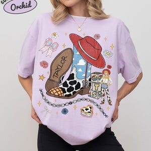 May include: An orchid-coloured t-shirt featuring a graphic print of a cowboy boot with a cow-print pattern, a red hat, a character, and alphabet blocks spelling TS. The text TAYLOR is on the boot sole, and a beaded string reads I KNEW IT.