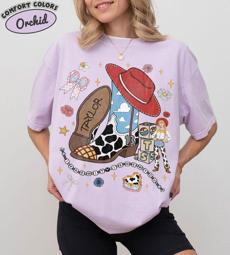 May include: An orchid-coloured t-shirt featuring a graphic print of a cowboy boot with a cow-print pattern, a red hat, a character, and alphabet blocks spelling TS. The text TAYLOR is on the boot sole, and a beaded string reads I KNEW IT.