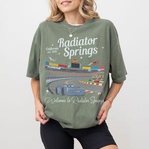 Puede incluir: Camiseta verde oliva con un gr&aacute;fico de Radiator Springs, California, con una pista de carreras y un coche azul. El texto "Radiator Springs" y "Welcome to Radiator Springs" est&aacute;n en la camiseta.