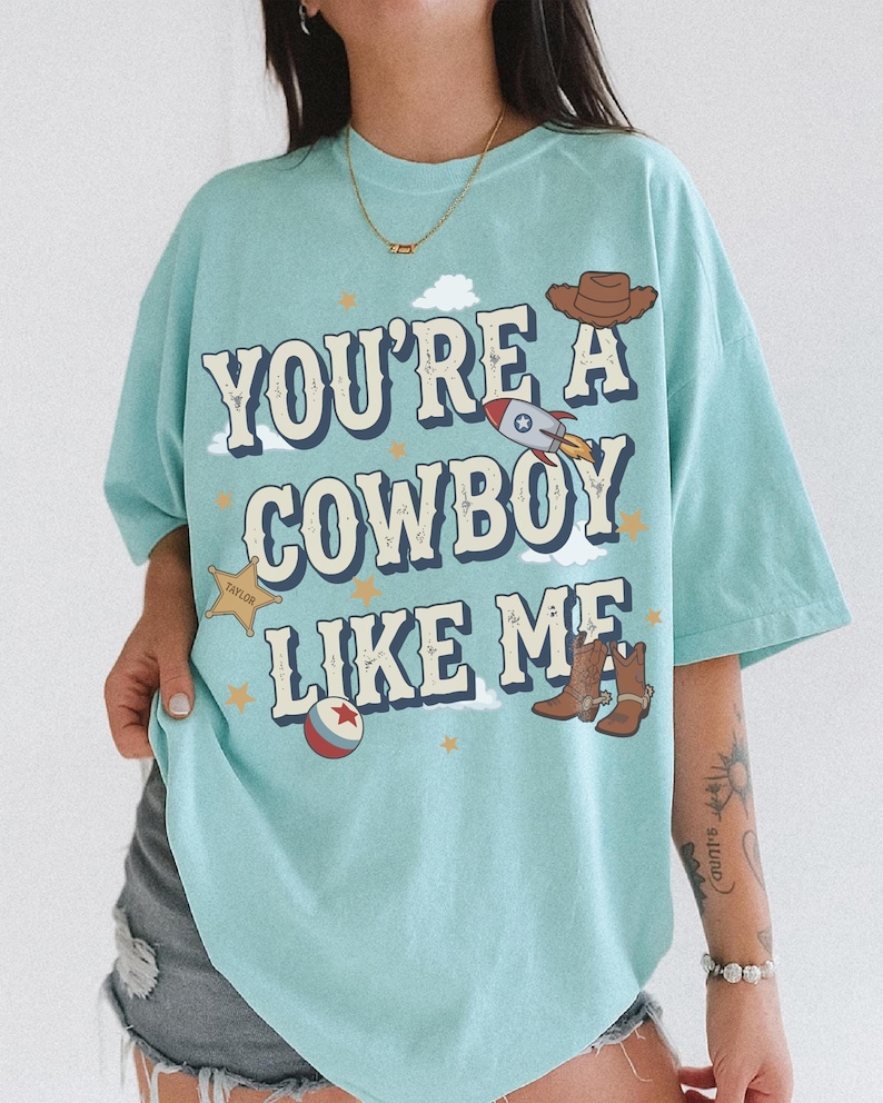 May include: A light blue t-shirt featuring the text "YOU'RE A COWBOY LIKE ME" in a dark blue font. The design includes a brown cowboy hat, a rocket ship, a yellow sheriff star badge labeled "TAYLOR", a red and white ball, and brown cowboy boots.
