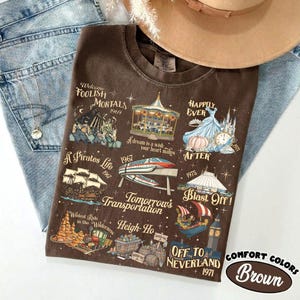 K&ouml;nnte beinhalten: Braunes T-Shirt mit Grafiken und Texten von Themenpark-Fahrgesch&auml;ften. Das Shirt enth&auml;lt S&auml;tze wie "Foolish Mortals", "A Pirate's Life" und "Off to Neverland". Das Shirt liegt auf einer blauen Jeans und neben einem beigen Hut.