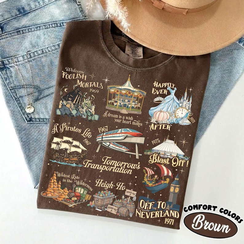 K&ouml;nnte beinhalten: Braunes T-Shirt mit Grafiken und Texten von Themenpark-Fahrgesch&auml;ften. Das Shirt enth&auml;lt S&auml;tze wie "Foolish Mortals", "A Pirate's Life" und "Off to Neverland". Das Shirt liegt auf einer blauen Jeans und neben einem beigen Hut.