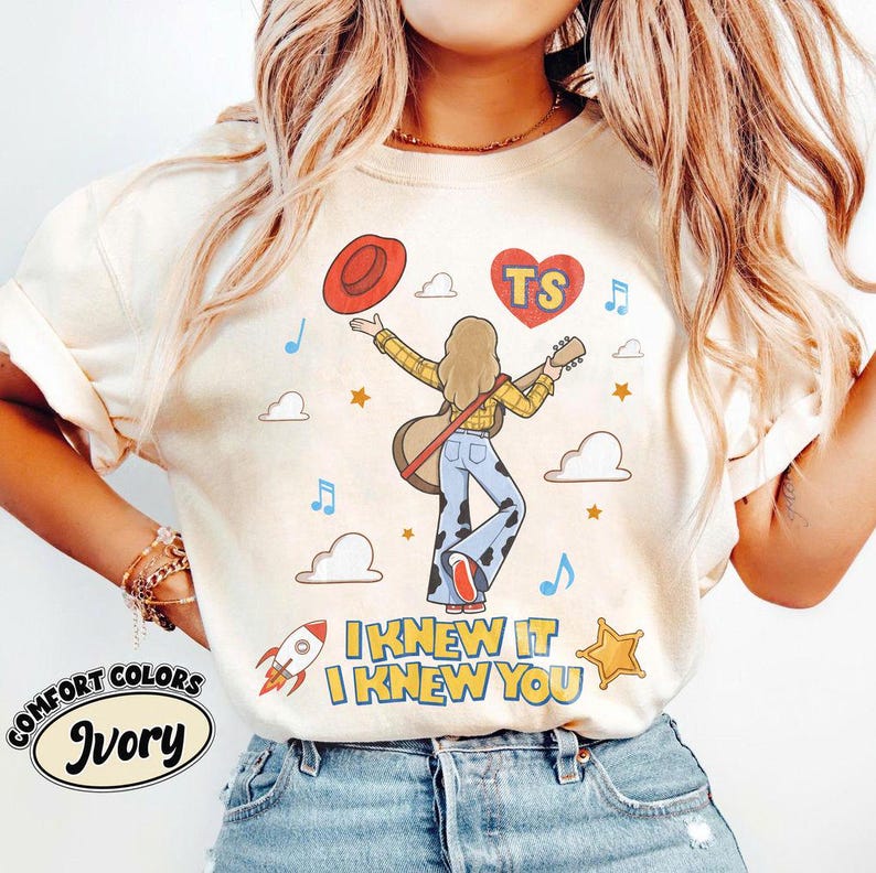 May include: An ivory t-shirt featuring a graphic of a person with long hair playing a guitar, wearing a yellow plaid shirt and blue cow-print bell-bottom trousers. The design includes a red hat, a heart with the text TS, clouds, stars, a rocket, and the text I KNEW IT I KNEW YOU.
