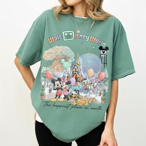 May include: Green t-shirt featuring a colorful graphic of Disney characters and landmarks, including Mickey and Minnie Mouse, Goofy, Donald and Daisy Duck. The text "Walt Disney World" and "The happiest place in earth" are also displayed.