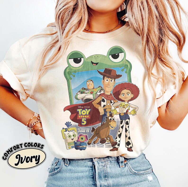 May include: An ivory t-shirt featuring a graphic print of Toy Story 5 characters including Woody, Jessie, Buzz Lightyear, Bullseye, and others framed by a green frog outline. The text reads Toy Story 5 In Theaters June 19.