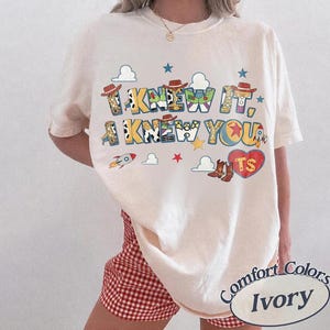 May include: An ivory short-sleeve t-shirt featuring a graphic print with the text I KNEW IT, I KNEW YOU. The design includes colorful cartoon elements like cowboy hats, a rocket, a red heart with the letters TS, and various cloud shapes.