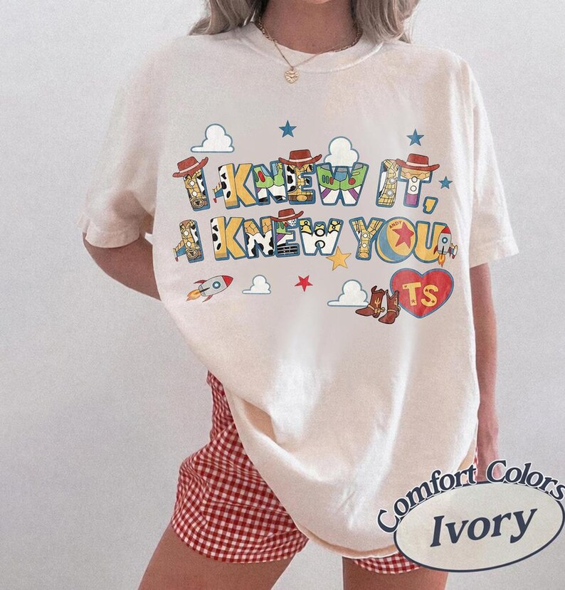 May include: An ivory short-sleeve t-shirt featuring a graphic print with the text I KNEW IT, I KNEW YOU. The design includes colorful cartoon elements like cowboy hats, a rocket, a red heart with the letters TS, and various cloud shapes.