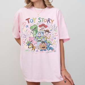 K&ouml;nnte beinhalten: Ein hellrosa T-Shirt mit dem Schriftzug "TOY STORY" in bunten Buchstaben. Das Shirt zeigt eine Grafik verschiedener Charaktere aus dem Animationsfilm, darunter Woody, Buzz Lightyear und Rex. Das Design ist von bunten Kritzeleien umgeben.