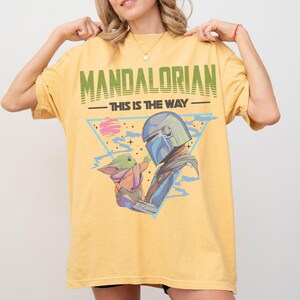 Może przedstawiać: Ż&oacute;łty t-shirt z nadrukiem graficznym przedstawiającym Mandalorianina i Grogu. Tekst głosi MANDALORIAN oraz THIS IS THE WAY. Wz&oacute;r zawiera niebieski tr&oacute;jkąt, r&oacute;żową planetę i gwiazdy. Koszulka ma kr&oacute;tkie rękawy i okrągły dekolt.
