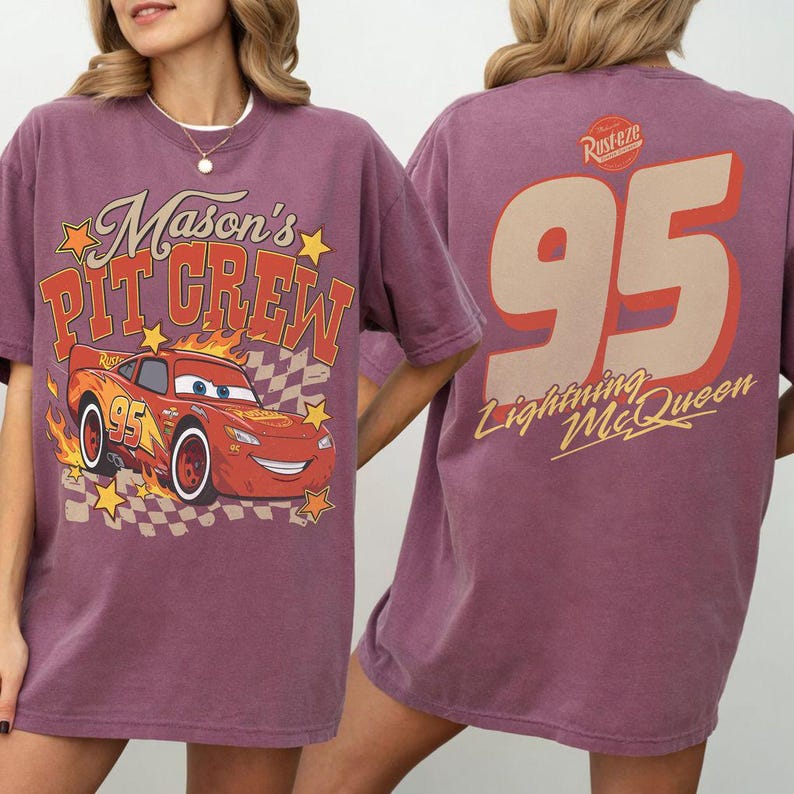 Op de afbeelding: Een mauve t-shirt met korte mouwen met een afbeelding op de voorkant van een rode raceauto met de tekst Mason's Pit Crew en gele sterren. De achterkant toont het nummer 95 in een groot beige lettertype met de tekst Rust-eze en Lightning McQueen.