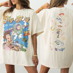 May include: Cream-colored t-shirt featuring a colorful graphic of Toy Story characters, including Buzz Lightyear and Woody, with the text "Toy Story Est. 1995." The back of the shirt displays additional character illustrations.