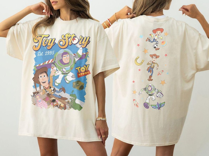 May include: Cream-colored t-shirt featuring a colorful graphic of Toy Story characters, including Buzz Lightyear and Woody, with the text "Toy Story Est. 1995." The back of the shirt displays additional character illustrations.