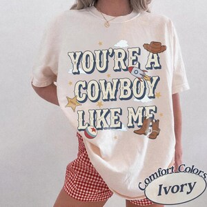 May include: An ivory t-shirt featuring the text "YOU'RE A COWBOY LIKE ME" in dark blue letters. The graphic includes a brown cowboy hat, a rocket ship, a sheriff star, a ball, and brown cowboy boots, accented with small yellow stars and clouds.