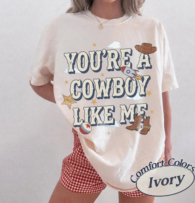 May include: An ivory t-shirt featuring the text "YOU'RE A COWBOY LIKE ME" in dark blue letters. The graphic includes a brown cowboy hat, a rocket ship, a sheriff star, a ball, and brown cowboy boots, accented with small yellow stars and clouds.