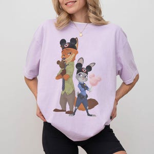 Pu&ograve; includere: T-shirt lilla chiaro con un'illustrazione a cartoni animati di Nick Wilde e Judy Hopps di Zootropolis. Nick, una volpe, indossa un cappello e tiene in mano un orsacchiotto. Judy, una coniglia, indossa un'uniforme da poliziotta e tiene lo zucchero filato.