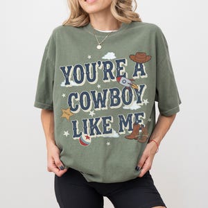 May include: An olive green short-sleeve t-shirt featuring the dark blue text "YOU'RE A COWBOY LIKE ME" with graphic illustrations of a brown cowboy hat, a rocket ship, a gold sheriff star, a pair of brown cowboy boots, and a red and blue ball.